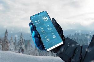 Smart phone in hand with glove with weather app on display. Winter, snowing time. Online service with weather forecast for several days concept photo