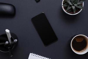Minimalist and organized work desk with a blank smart phone surrounded by a plant, pens, coffee and notepad. Blank screen for mockup app presentation. Office work desk composition, top view, flat lay photo
