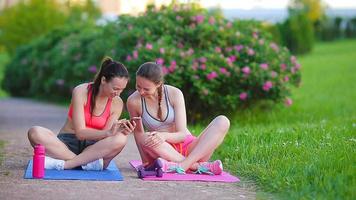 jóvenes mujeres activas de fitness haciendo ejercicios al aire libre y mirando el teléfono inteligente. dos chicas deportivas practican deportes en narure en el parque video