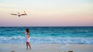 aanbiddelijk weinig meisje met vliegend vlieger Aan tropisch strand. kind Speel Aan oceaan kust met mooi zonsondergang. langzaam beweging. video