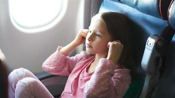 Adorable little girl traveling by an airplane. Cute kid listening music near window in aircraft