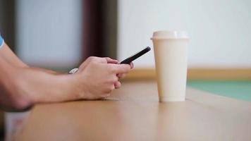 primer plano de manos masculinas sosteniendo un teléfono celular y un vaso de café en la cafetería. hombre usando teléfono inteligente móvil. niño tocando una pantalla de su smarthone. fondo borroso, horizontal. video