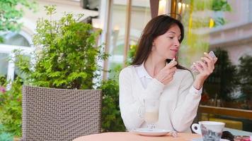 belle femme peignant ses lèvres avec du rouge à lèvres assis dans un café en plein air. jeune fille avec un délicieux cappuccino sucré. video