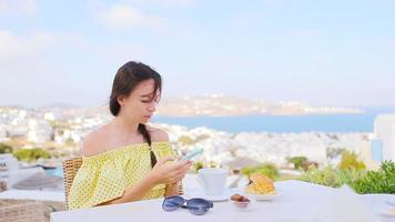 Caucasian girl on breakfast with smartphone at outdoor cafe with amazing view on Mykonos town. video