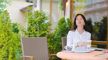 Porträt einer schönen Frau, die im Café im Freien sitzt, Kaffee trinkt und Smartphone benutzt. junges mädchen, das instagram sieht und köstlichen süßen cappuccino trinkt. video