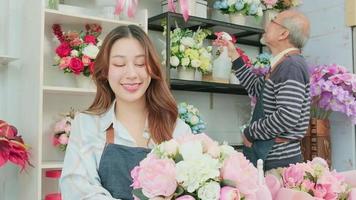 jong mooi Aziatisch vrouw bloemist ondernemer regelen van een bundel van bloesems werken met oud Mens winkelier, gelukkig werk in kleurrijk bloem winkel op te slaan met vers bloeit, klein bedrijf, familie MKB video