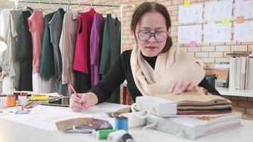 designer de moda feminina asiática de meia-idade trabalhando em um estúdio por meio de ideias desenhando esboços com fios coloridos e costurando para uma coleção de design de vestidos, alfaiate profissional empresária de sme. video