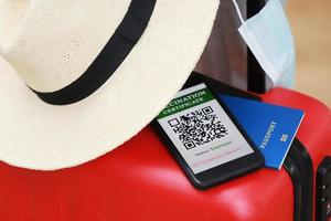 New normal concept. Close up view of a smartphone with an immune digital health passport, a red suitcase, protective mask, straw hat, passport and sunglasses. Traveling by plane during a pandemic. photo