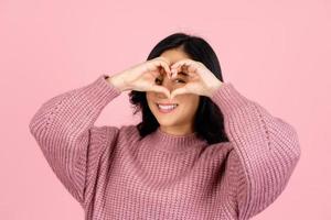 Portrait of attractive cheerful asian young woman showing heart shape looking eye sindow spy isolated over pink pastel color background photo