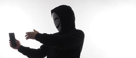 hacker anónimo y mascarilla con smartphone en la mano. hombre con camisa de capucha negra sosteniendo y usando un teléfono móvil con fondo blanco. representan el concepto de piratería informática o robo de datos personales foto