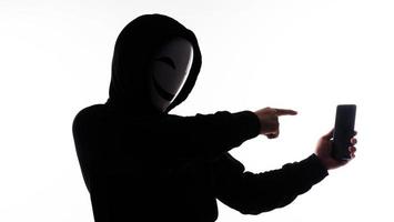 Hacker Anonymous and face mask with smartphone in hand. Man in black hood shirt holding and using mobile phone on white background. Represent cyber crime data hacking or stealing personal data concept photo