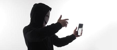 hacker anónimo y mascarilla con smartphone en la mano. hombre con camisa de capucha negra sosteniendo y usando un teléfono móvil con fondo blanco. representan el concepto de piratería informática o robo de datos personales foto