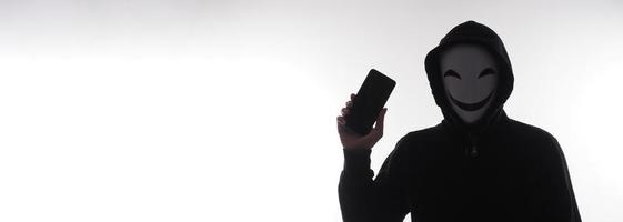 Hacker Anonymous and face mask with smartphone in hand. Man in black hood shirt holding and using mobile phone on white background. Represent cyber crime data hacking or stealing personal data concept photo