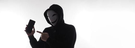 hacker anónimo y mascarilla con smartphone en la mano. hombre con camisa de capucha negra sosteniendo y usando un teléfono móvil con fondo blanco. representan el concepto de piratería informática o robo de datos personales foto