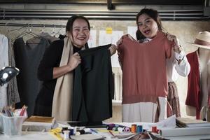 Asian middle-aged female fashion designer and young teen trainee tailor proud and cheerful with garments design work success, showing dresses job in studio, happy costume boutique small business. photo