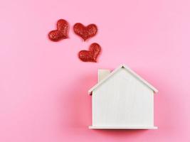 endecha plana de casa modelo de madera con corazones de brillo rojo sobre fondo rosa. casa de ensueño, hogar de amor, relación fuerte, san valentín. foto