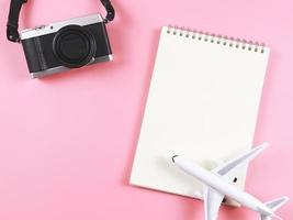 endecha plana de cuaderno abierto de página en blanco, modelo de avión y cámara sobre fondo rosa con espacio de copia. foto