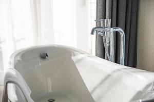 Chrome faucets and mixer on white ceramic bathtub in a retro style white bathroom in luxury home close to white curtain under sunlight. Idyllic bathroom interior photo