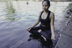 Asian woman practicing yoga and meditates near swimming pool outdoor. Fitness Asian lady sitting in lotus pose with closed eyes. photo