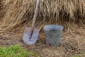 An iron shovel with a wooden handle and an iron bucket stand next to a haystack. photo