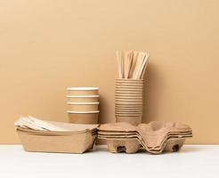 stack of brown disposable paper cups and a tray on a white table, wooden forks and knives, brown background photo