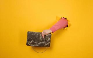 female hand is holding a small golden clutch bag with a metal chain on a yellow background. Part of the body sticking out of a torn hole in a paper background photo