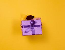 female hand holds a box with a gift on a yellow background, part of the body sticks out of a torn hole in a paper background. Congratulation, holiday surprise photo