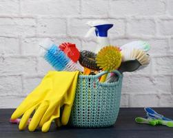 sponges, plastic brushes and bottles of detergents on a blue wooden table. Household cleaning items on white brick wall background photo