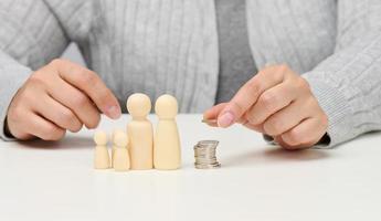 la mano femenina pone monedas en una pila y figuritas de madera de una familia sobre una mesa blanca. concepto de economía e inversión, planificación presupuestaria. recibir subsidios del estado, impuestos foto