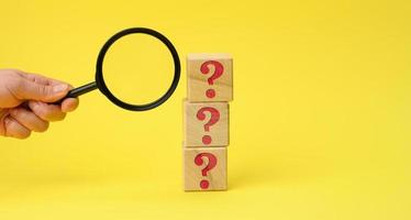 wooden cubes with question marks and a female hand holds a magnifying glass on a yellow background. The concept of finding answers to unknown questions photo