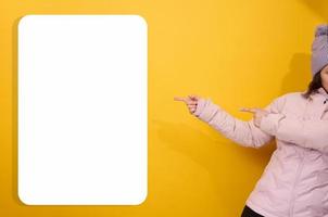 woman in a winter pink jacket and hat shows with her hands to the side on a blank white billboard on a yellow background. Place for announcements, advertisements. photo