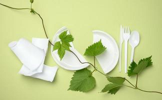 plato de plástico blanco roto, tenedor, cuchillo y rama con hojas verdes sobre un fondo verde. el concepto de evitar el plástico, preservar el medio ambiente foto