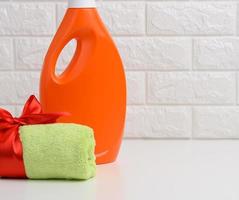 una toalla verde enrollada atada con una cinta de seda roja y una botella de plástico naranja de detergente líquido para ropa en un estante blanco en el baño foto