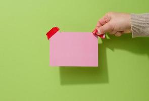 female hand holding blank pink sheet of paper with red sticky tape on green background photo