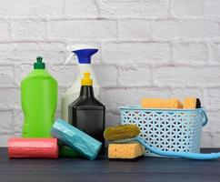 esponjas, cepillos de plástico y botellas de detergentes sobre una mesa de madera azul. artículos de limpieza del hogar sobre fondo de pared de ladrillo blanco foto