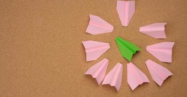 green paper airplane surrounded in a ring by pink paper airplanes on a brown background photo