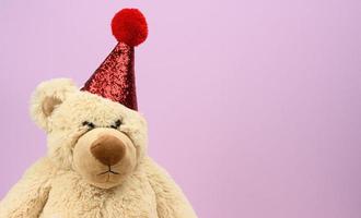 triste oso de peluche beige con una gorra roja se sienta sobre un fondo morado foto