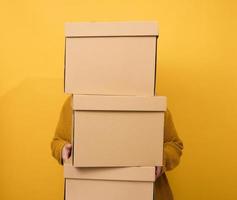 a woman in a sweater holds a stack of brown cardboard boxes on a yellow background. Relocation photo