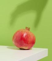 la granada roja entera y madura se encuentra sobre una mesa blanca, la sombra de la mano alcanza la fruta. fondo verde