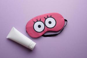 white plastic tube for cosmetics and pink textile sleep mask on purple background photo