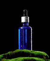 botella de vidrio azul con soportes de pipeta sobre musgo verde, fondo negro. maqueta de marca de spa de cosméticos foto