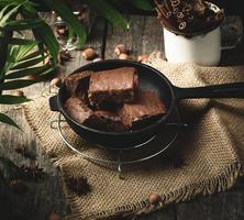 baked pieces of chocolate brownie cake with walnuts in a black metal frying pan on a wooden table, top view photo