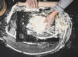 two female hands knead the dough from white wheat flour on a black table, top view photo