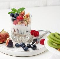 transparent glass with granola poured with yogurt, on top of ripe raspberries, blueberries and figs on a white table. Healthy breakfast photo