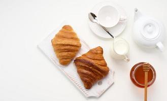 baked croissants, white ceramic teapot and empty cup and saucer, jar of honey on a white table, top view photo