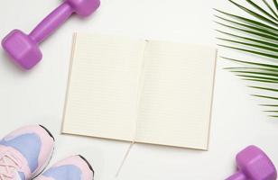 abra un cuaderno en blanco, un par de zapatos deportivos de gimnasia y pesas sobre un fondo blanco. lugar para grabar, vista superior foto