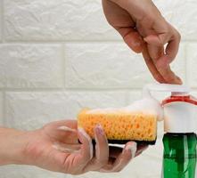 female hand holds kitchen sponge with white foam near plastic dispenser, other hand squeezes out liquid photo