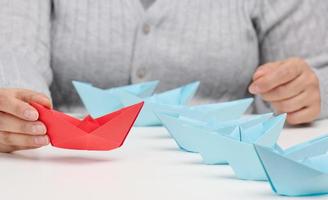 group of blue paper boats follows a res boat in front of a white table. The concept of a strong and charismatic leader in a team photo