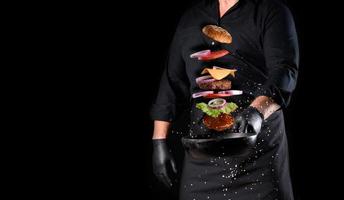 hombre con uniforme negro sosteniendo una sartén redonda de hierro fundido con ingredientes de hamburguesa con queso levitando foto