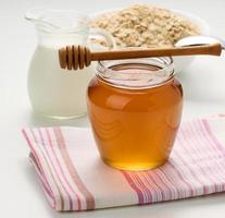 honey in a glass transparent jar and a wooden stick on a white table, behind a decanter with milk and oatmeal photo